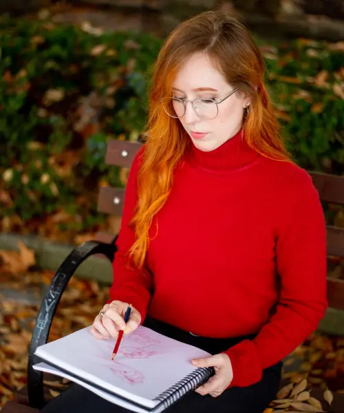 young female artist with red hair and glasses looking down and drawing